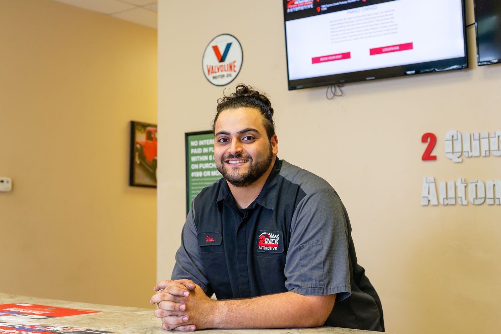 A smiling professional in a uniform stands behind a service desk in an auto shop with a Valvoline sign on the wall. | 2 QuickAuto, Car & Tire Repair | Copperfield