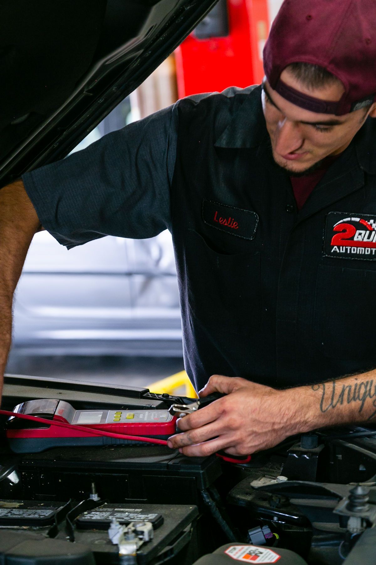 A mechanic wearing a maroon cap and dark uniform uses a red multimeter to test a car battery under an open hood. | 2 QuickAuto, Car & Tire Repair | Copperfield