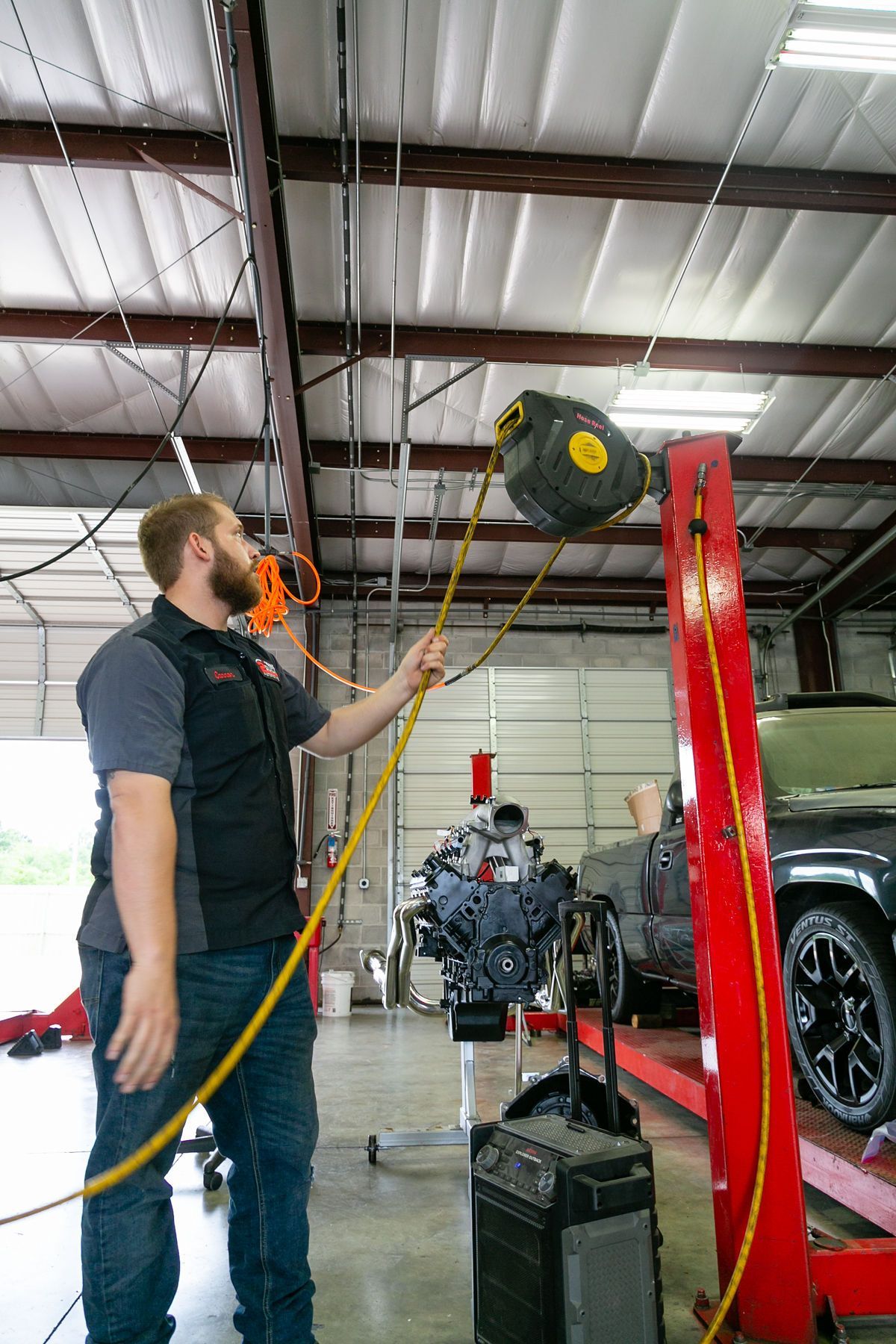 A person in a mechanic shop pulls down a yellow extension cord from a ceiling-mounted reel near a vehicle on a red lift. | 2 QuickAuto, Car & Tire Repair | Copperfield