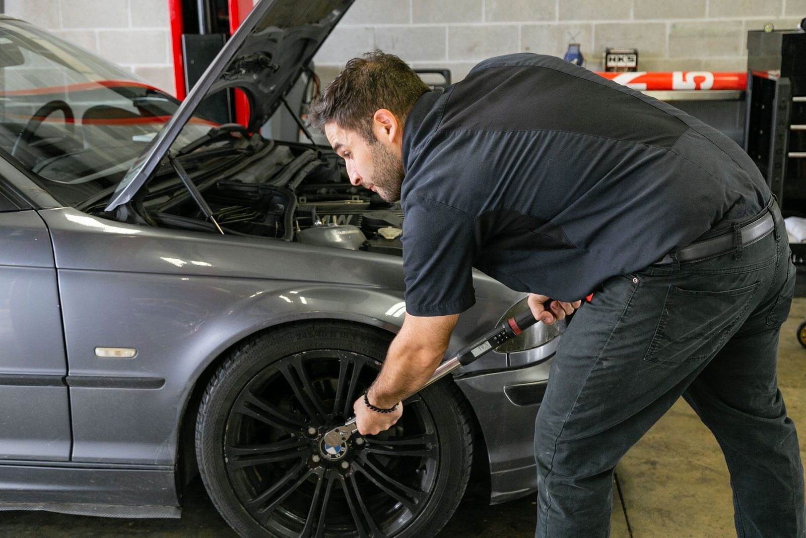 A person in a garage uses a torque wrench to tighten a lug nut on the wheel of a gray car with its hood open. | 2 QuickAuto, Car & Tire Repair | Copperfield