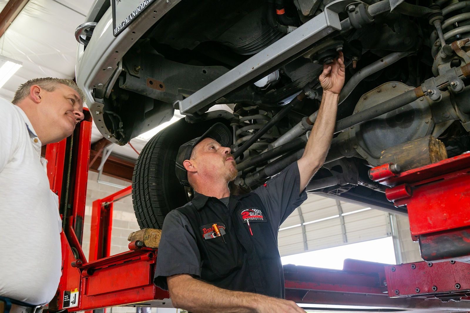 A mechanic points to a car component on a lift while explaining it to another person in a repair shop. | 2 QuickAuto, Car & Tire Repair | Copperfield