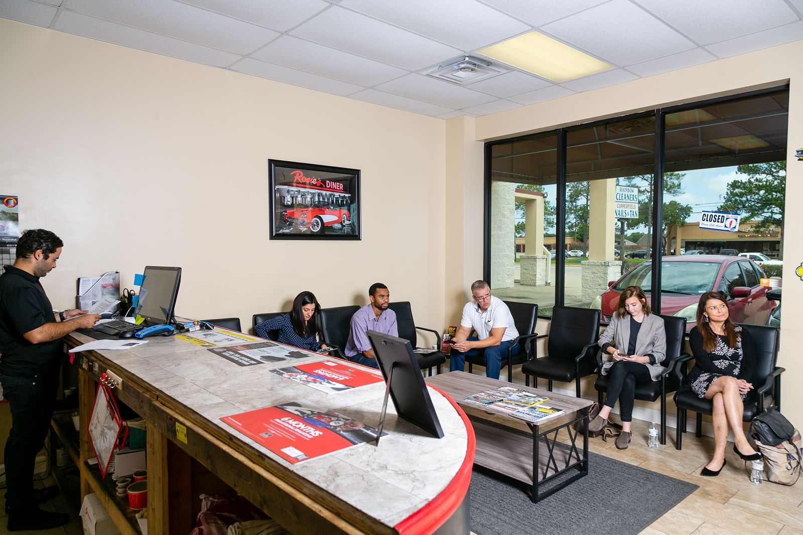 A person behind a desk assists customers waiting in a brightly lit, modern office lobby with large windows. | 2 QuickAuto, Car & Tire Repair | Copperfield