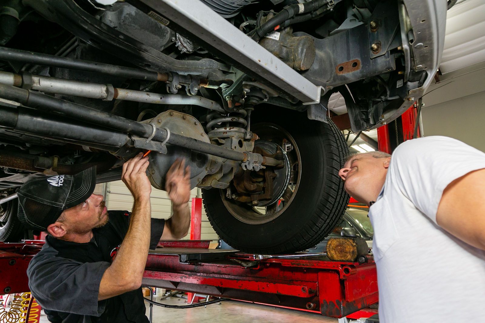 Two mechanics inspect the underside of a vehicle raised on a red automotive lift in a workshop. | 2 QuickAuto, Car & Tire Repair | Copperfield
