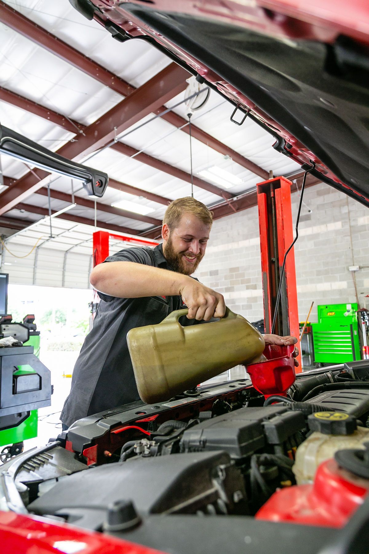 A technician smiling as they pour oil into the engine of a red vehicle inside a workshop.  | 2 QuickAuto, Car & Tire Repair | Copperfield