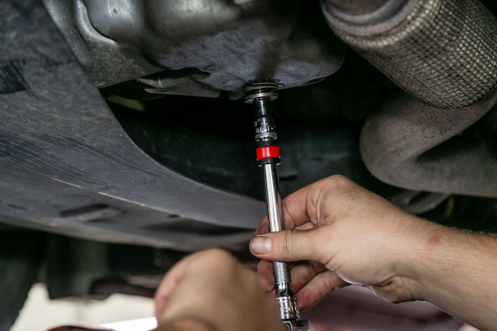 Hands using a socket wrench to remove the drain plug from the underside of a car engine. | 2 QuickAuto, Car & Tire Repair | Copperfield