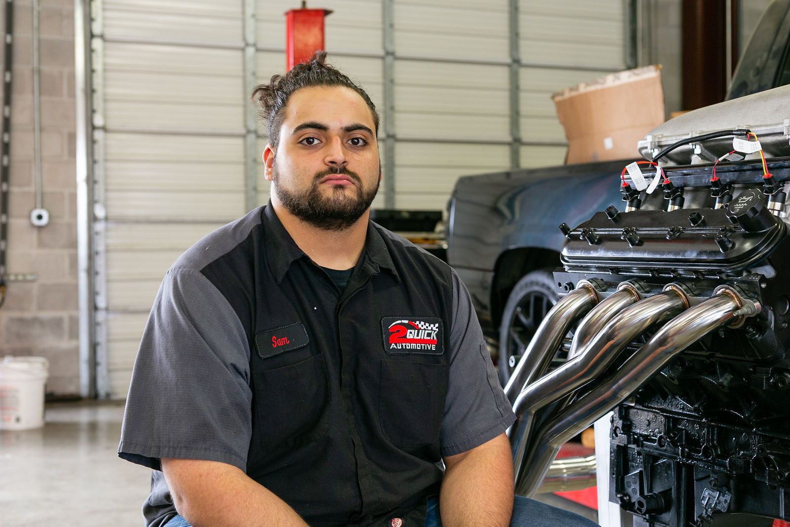 A mechanic sits in a garage next to a truck engine with metallic exhaust headers. | 2 QuickAuto, Car & Tire Repair | Copperfield