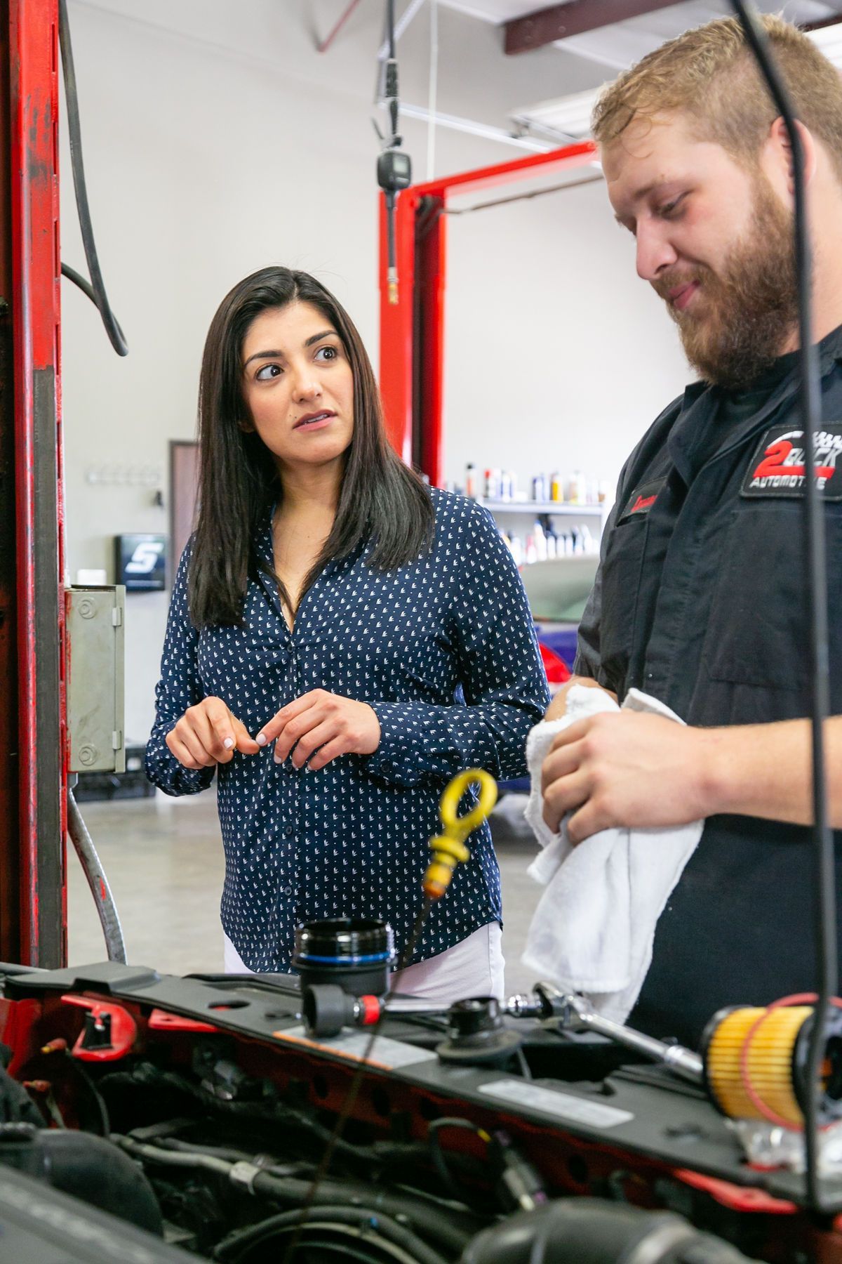 A mechanic and a customer stand in a garage, discussing the open hood of a car. | 2 QuickAuto, Car & Tire Repair | Copperfield