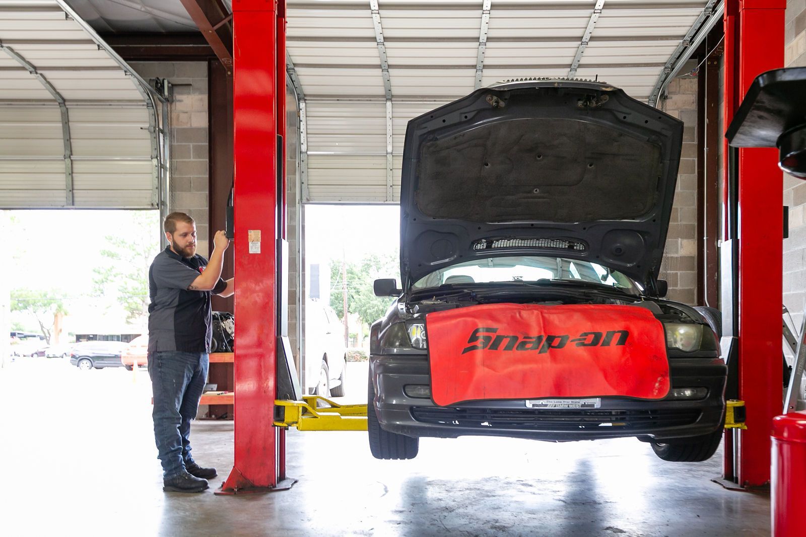 A mechanic standing by a car lifted on a hydraulic rack inside a garage with its hood open and a red fender cover. | 2 QuickAuto, Car & Tire Repair | Copperfield