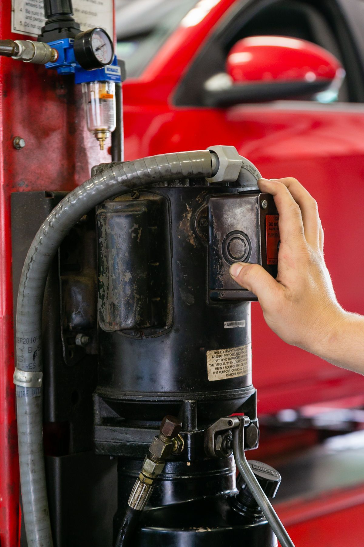 A person’s hand presses the red power button on a black motor unit in an auto repair shop with a red vehicle in the back. | 2 QuickAuto, Car & Tire Repair | Copperfield