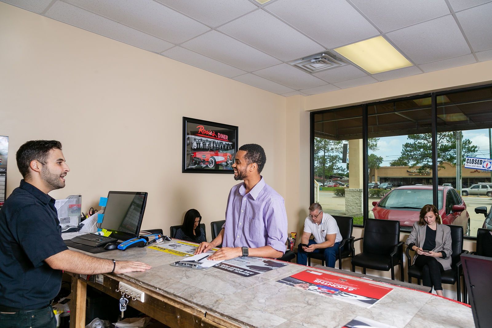 A customer stands at a service counter in a business office, speaking with an employee while two others wait nearby. | 2 QuickAuto, Car & Tire Repair | Copperfield