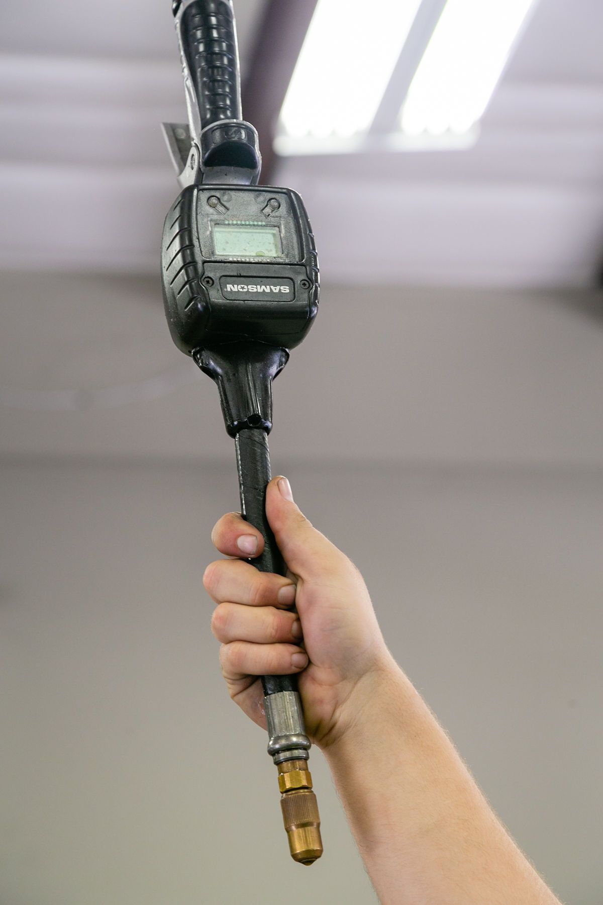 A hand holds an industrial oil control nozzle with a digital meter, hanging vertically against a blurred background. | 2 QuickAuto, Car & Tire Repair | Copperfield