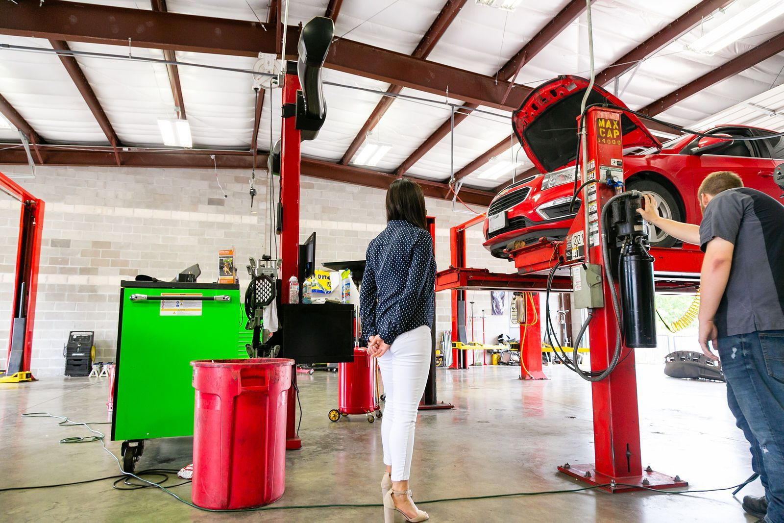 A technician and a woman observe a red car lifted on a mechanical hoist inside a well-lit automotive service center. | 2 QuickAuto, Car & Tire Repair | Copperfield
