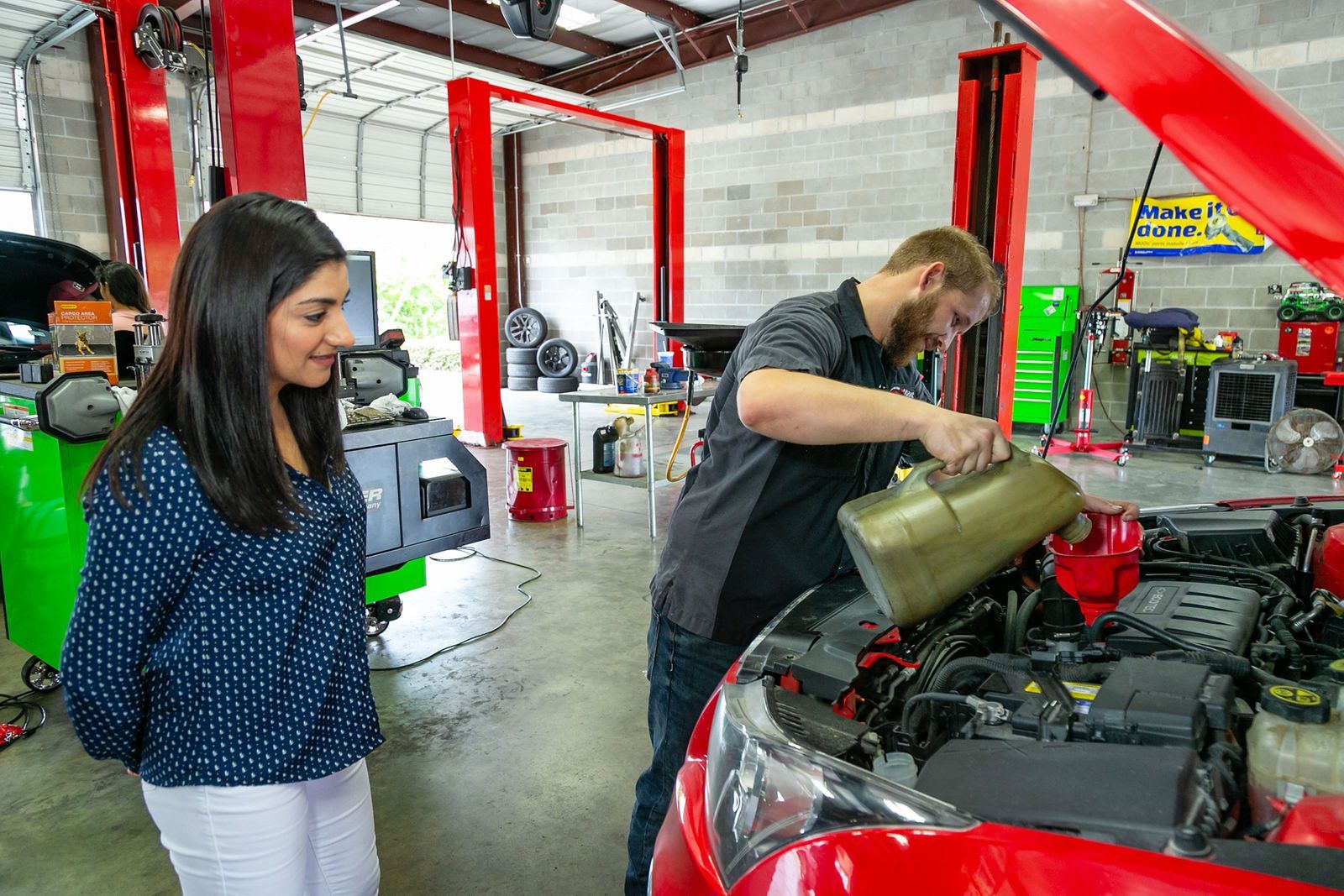 A mechanic pours oil into a car engine while a person watches in a well-lit auto repair shop with red vehicle lifts. | 2 QuickAuto, Car & Tire Repair | Copperfield