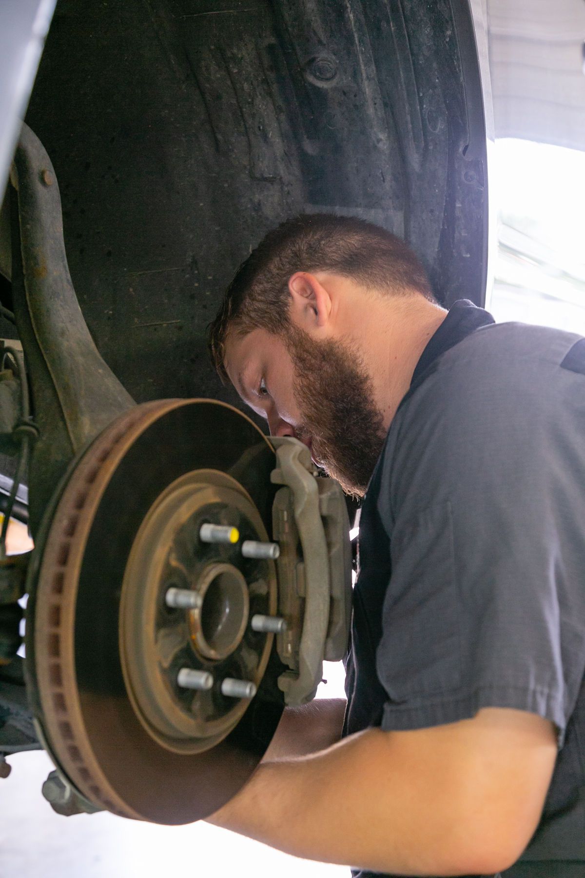 A mechanic with a beard wearing a grey shirt works on the disc brakes of a vehicle wheel assembly. | 2 QuickAuto, Car & Tire Repair | Copperfield