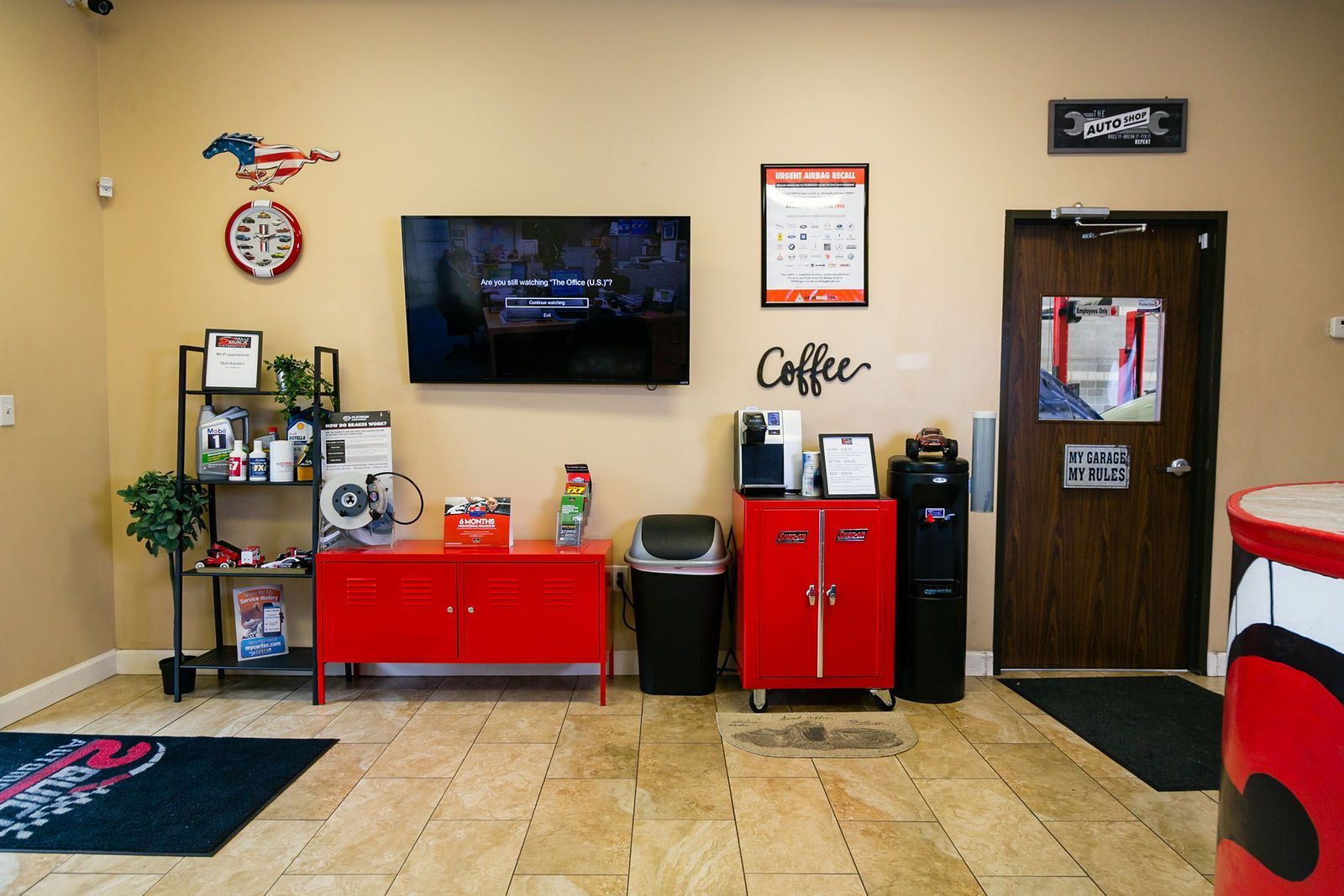 A waiting room with a TV, red cabinets, coffee station, shelf with supplies, and a doorway on a tiled floor. | 2 QuickAuto, Car & Tire Repair | Copperfield