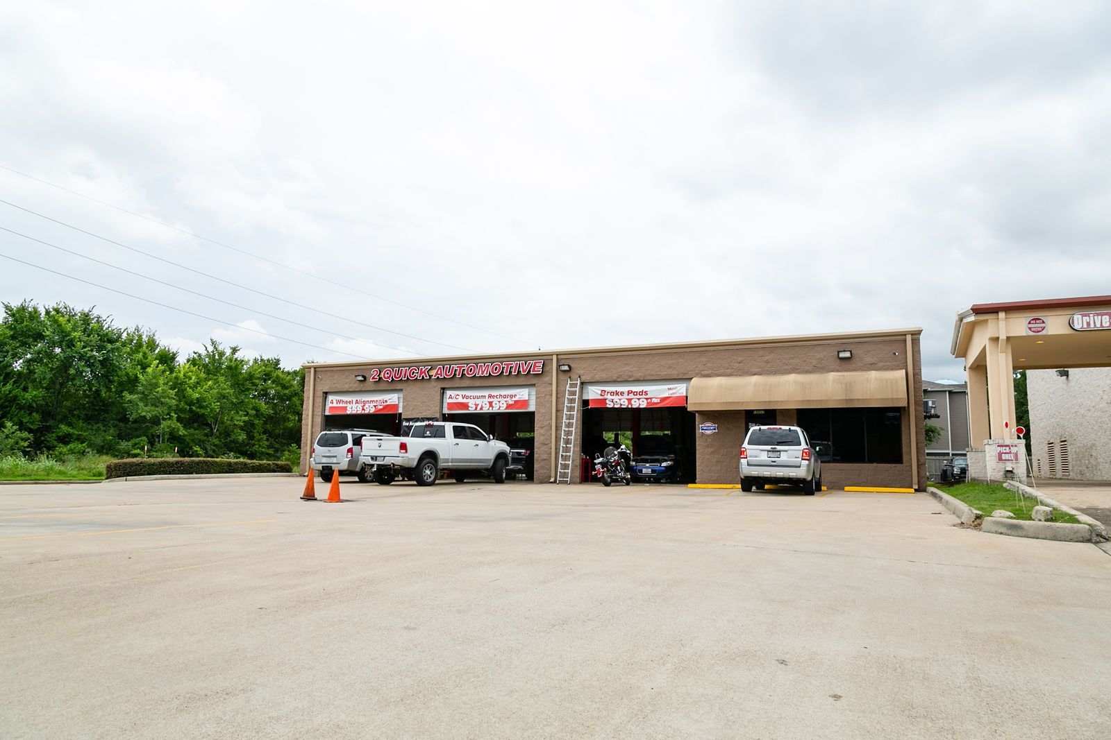 A tan brick automotive repair shop with multiple service bays, parked cars, and an adjacent structure under a cloudy sky. | 2 QuickAuto, Car & Tire Repair | Copperfield