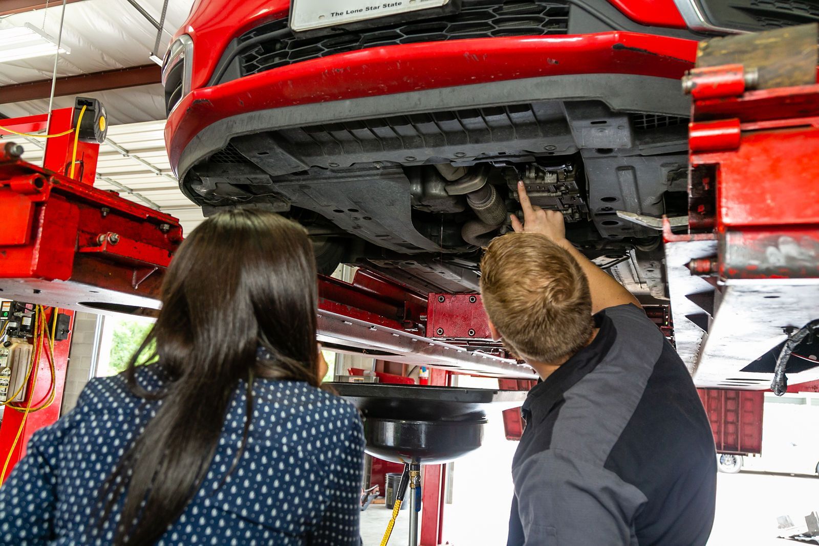 A technician points to the undercarriage of a red car raised on a lift while an observer watches in an auto shop. | 2 QuickAuto, Car & Tire Repair | Copperfield