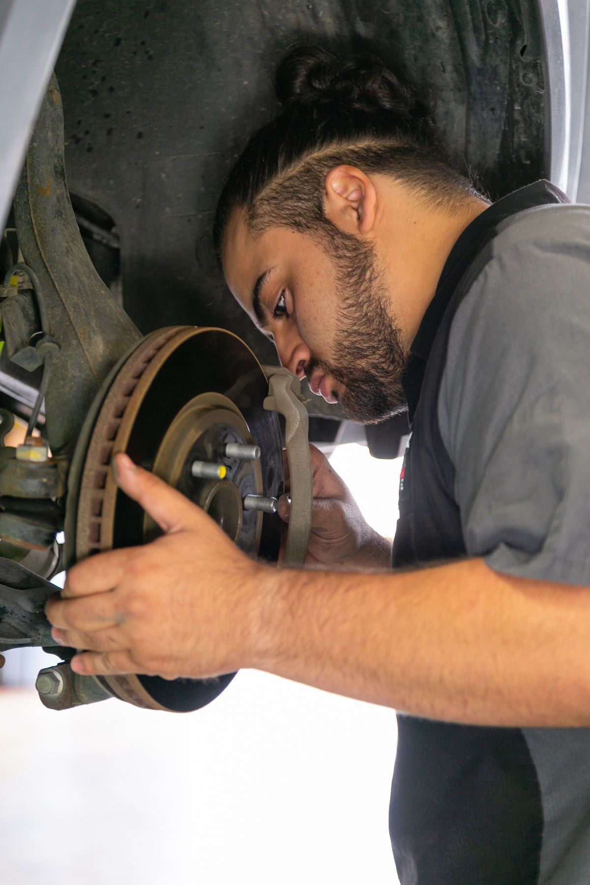 A mechanic in a grey uniform inspects a car’s brake disc assembly in a well-lit workshop. | 2 QuickAuto, Car & Tire Repair | Copperfield