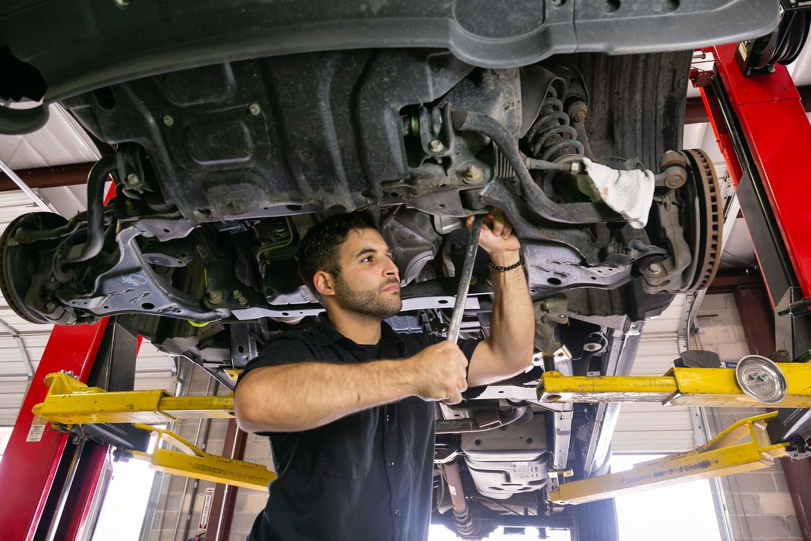 A mechanic in a black shirt uses a wrench to work on the underside of a vehicle lifted on a red and yellow auto lift. | 2 QuickAuto, Car & Tire Repair
