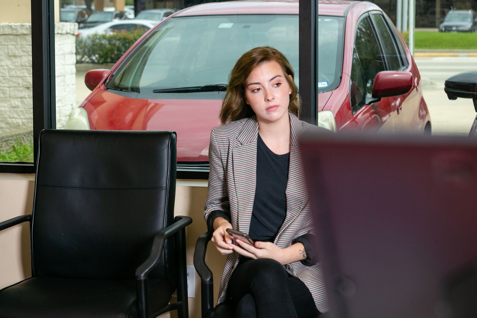 A person sits in a black chair, looking away while holding a phone, with a red car visible through a window behind them. | 2 QuickAuto, Car & Tire Repair | Copperfield