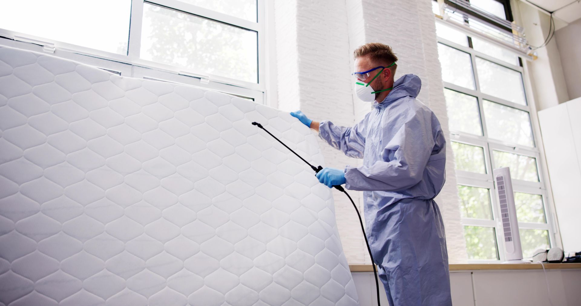 Person in protective suit spraying a mattress indoors near a window.