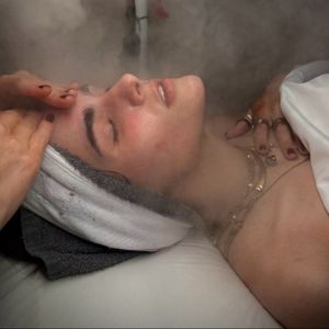 Person receiving facial steam treatment, eyes closed, hands on face, spa setting.