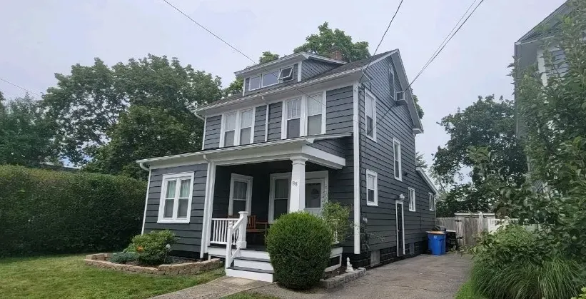 A large gray house with a white porch and a blue trash can in front of it.