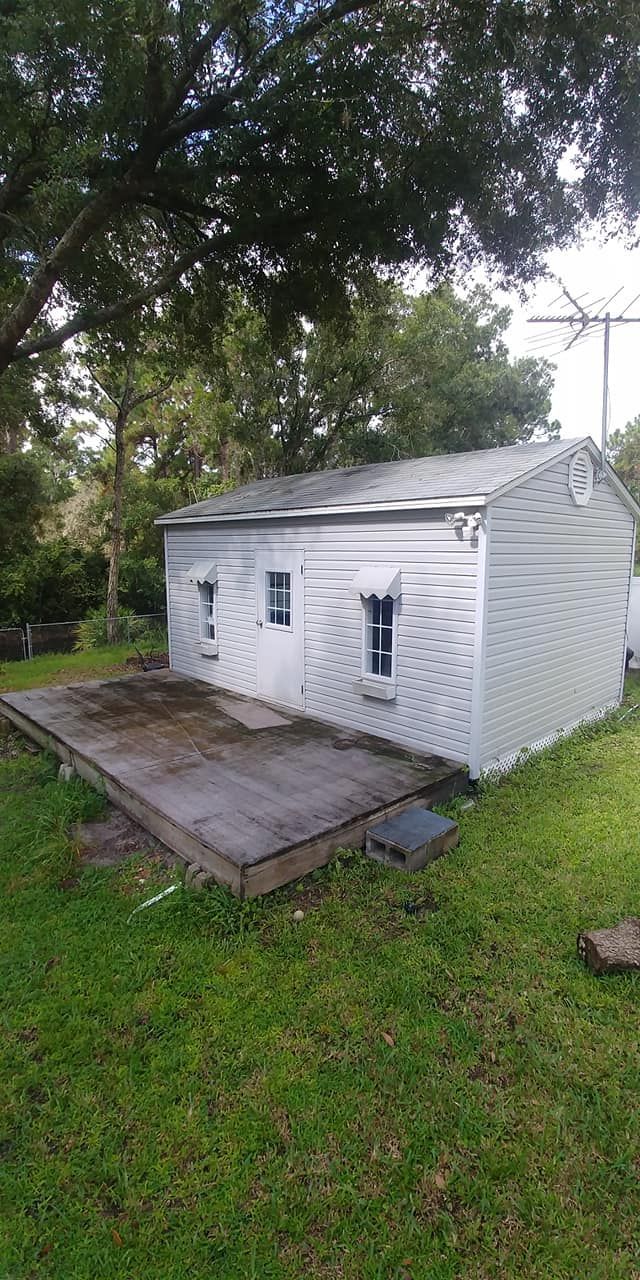 White shed with two small windows, set on a grassy lawn under a tree.