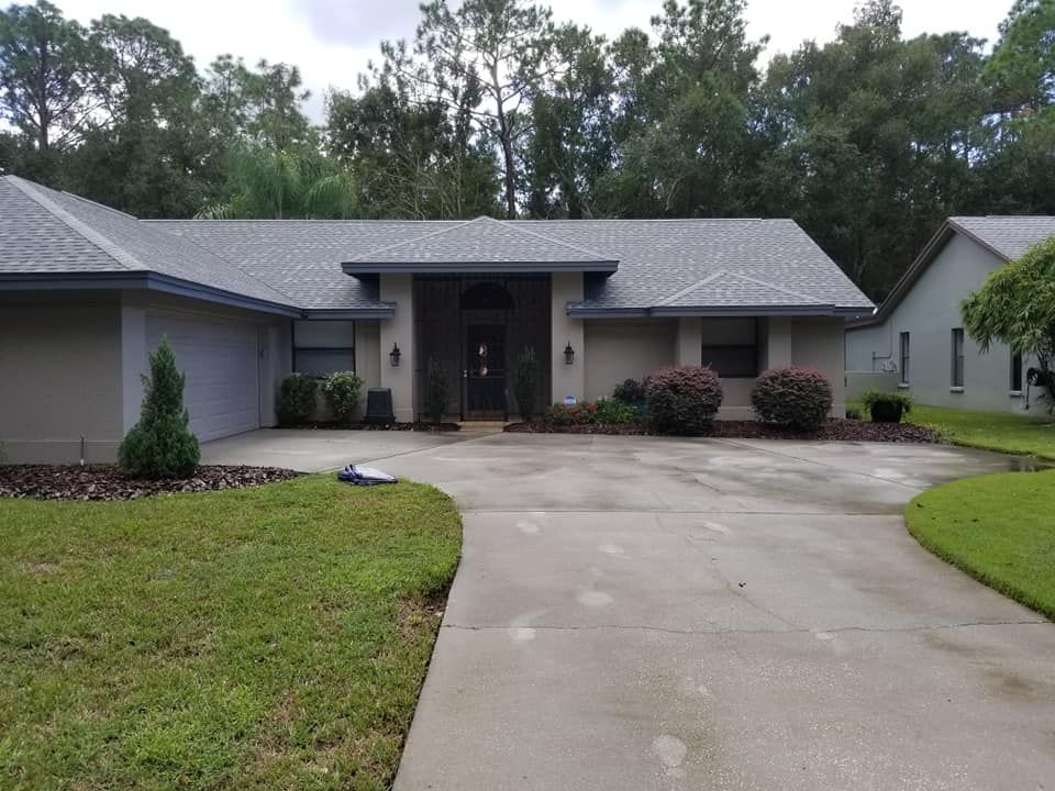 A single-story beige house with a gray roof, surrounded by green grass and a concrete driveway.