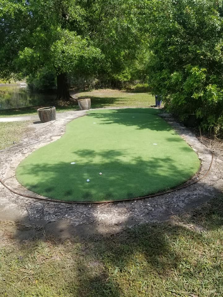 A putting green outdoors, with white golf balls on the green, surrounded by trees and a concrete border.