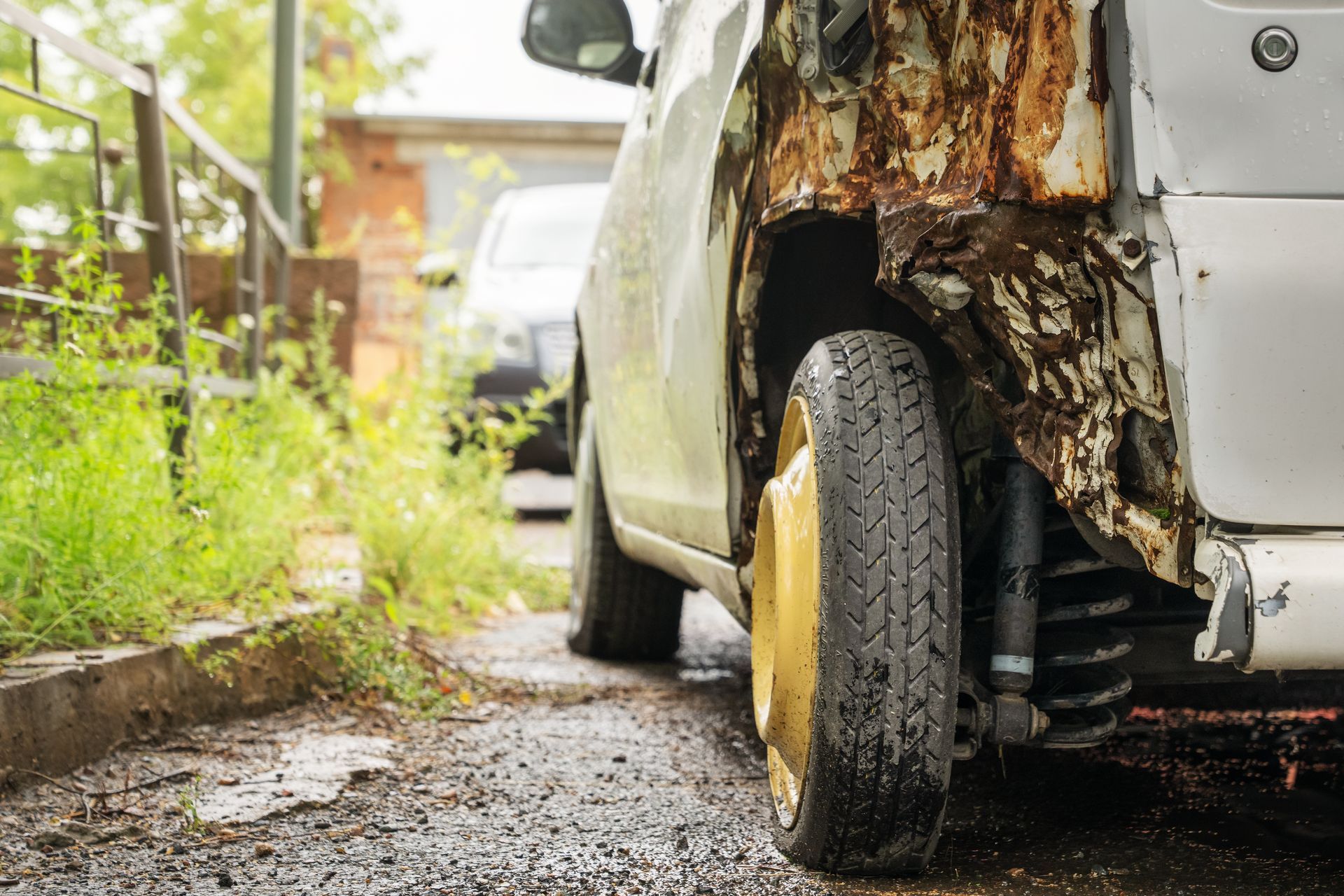 A damaged, rusted car with a visible tire parked on a gravel path near green vegetation. A damaged, rusted car with a visible tire parked on a gravel path near green vegetation.
