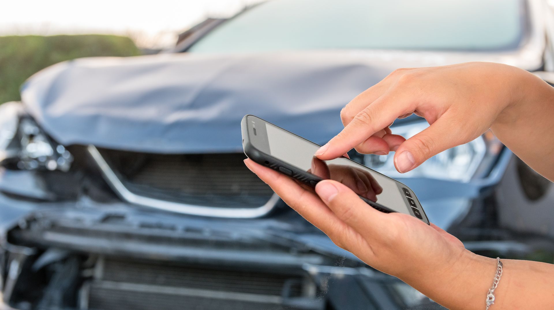 A person uses a smartphone in front of a car with a damaged front bumper and hood. A person uses a smartphone in front of a car with a damaged front bumper and hood.