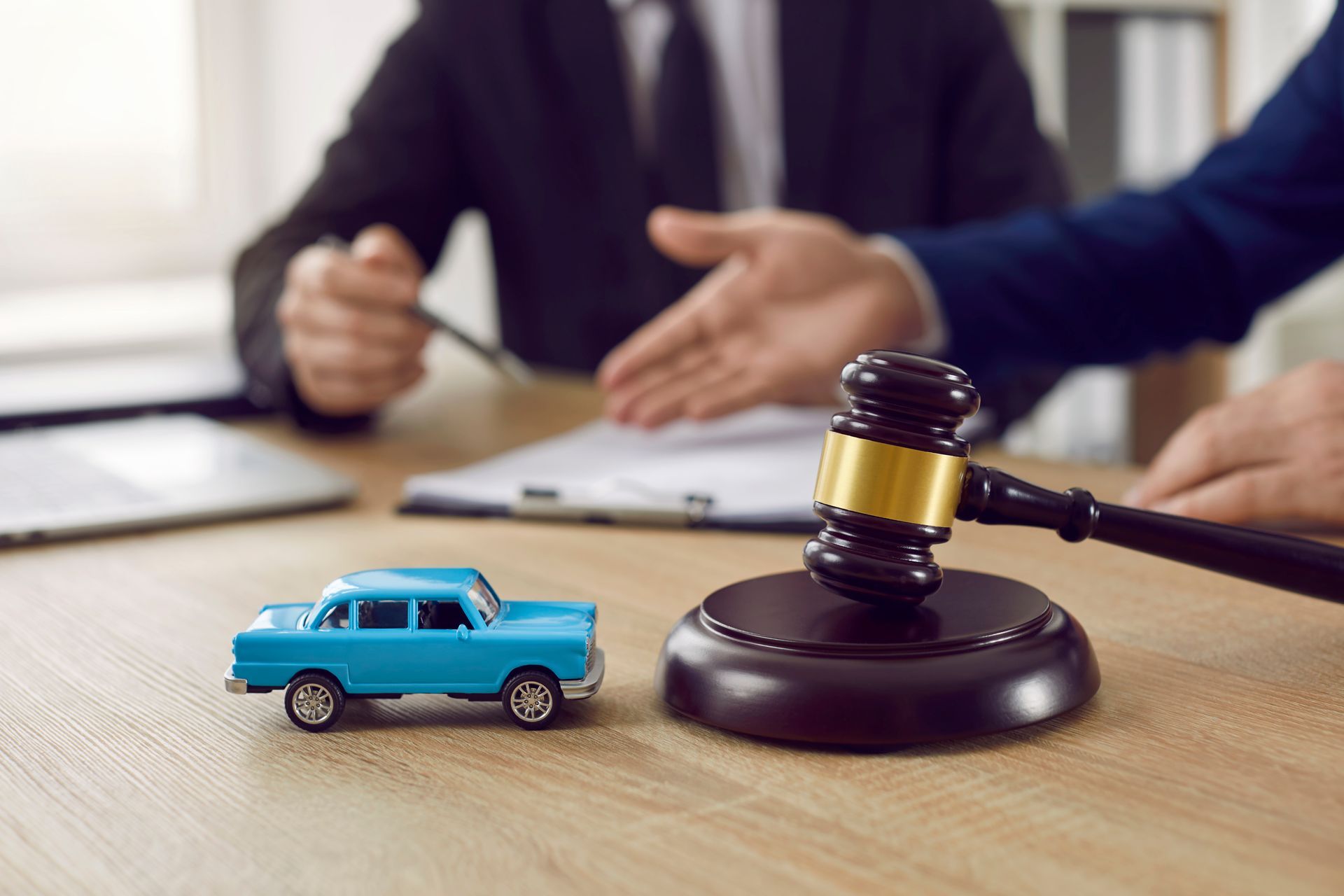 A blue toy car and a wooden judge’s gavel sit on a desk in front of two professionals in suits during a meeting. A blue toy car and a wooden judge’s gavel sit on a desk in front of two professionals in suits during a meeting.