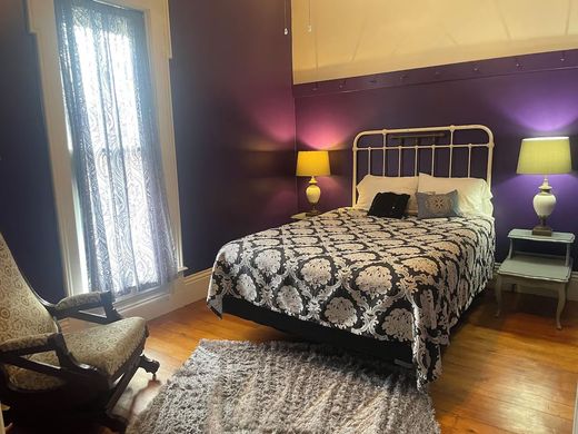 A bedroom with purple walls , a bed , two lamps , a rocking chair and a rug.