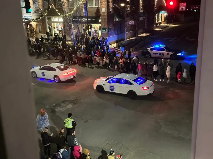Two police cars with flashing blue lights drive through a town square lined with spectators at night.