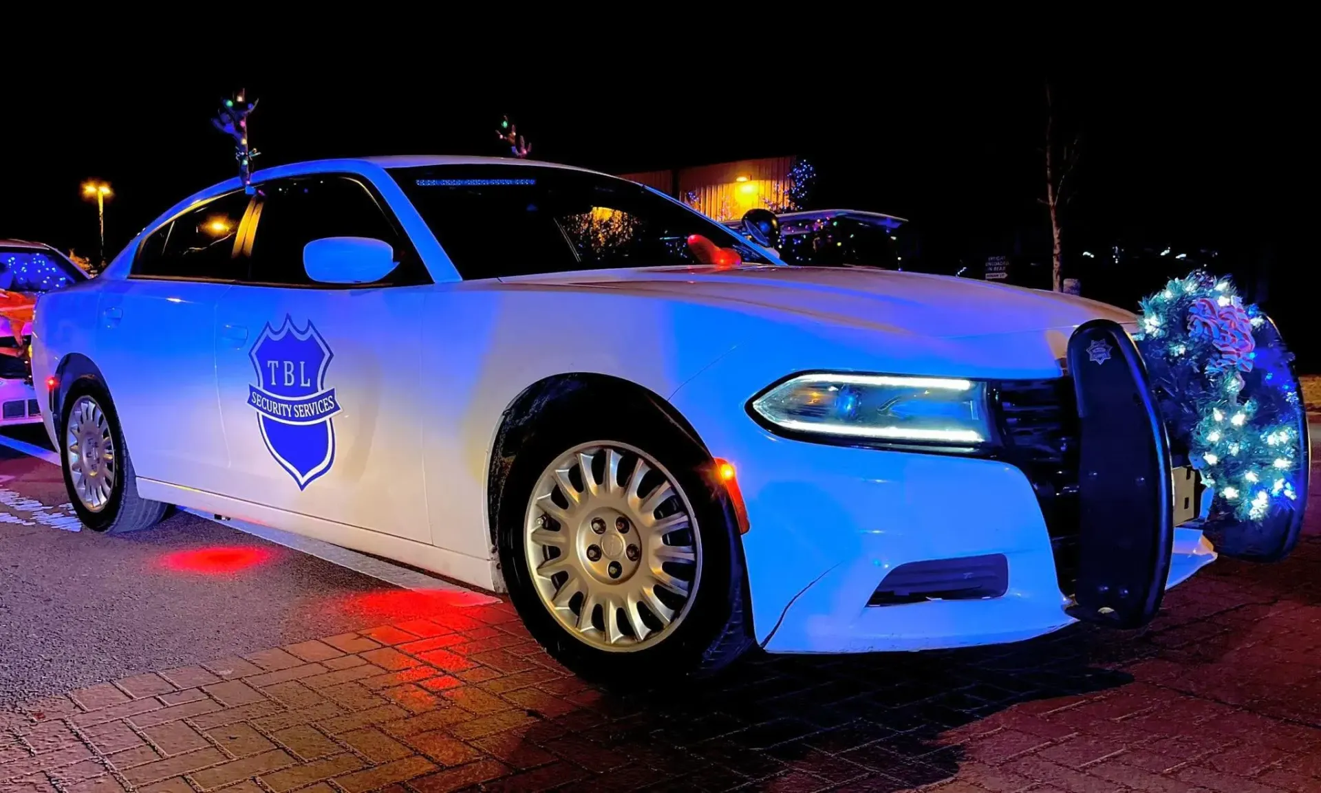 A white patrol car with blue police emblem and Christmas lights on the front bumper parked at night.
