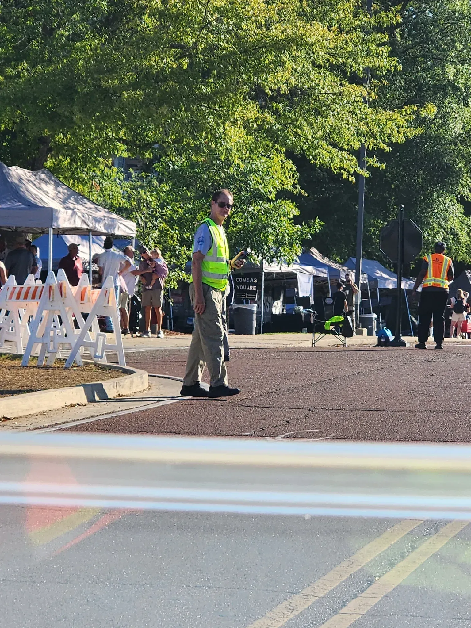 A person in a high-visibility vest and sunglasses stands at a public event with tents and crowd control barriers nearby.