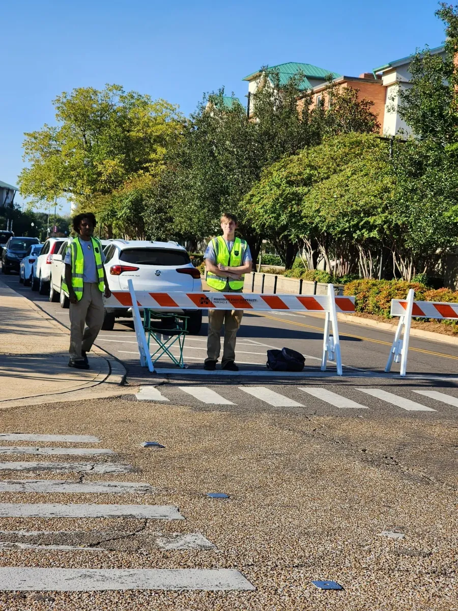 Two individuals in neon safety vests stand behind a road barricade blocking a street near a crosswalk.