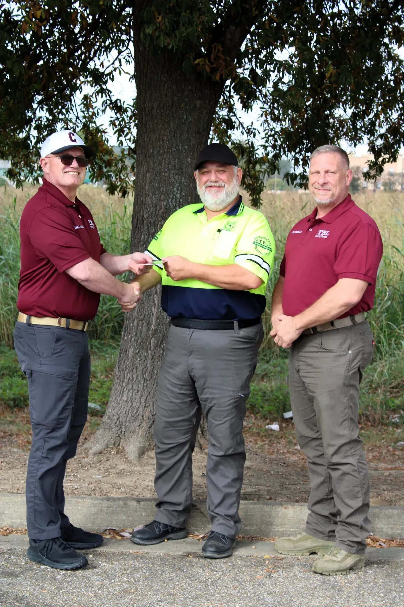 Three people stand outdoors near a tree, with the person on the left shaking hands with the person in the center.