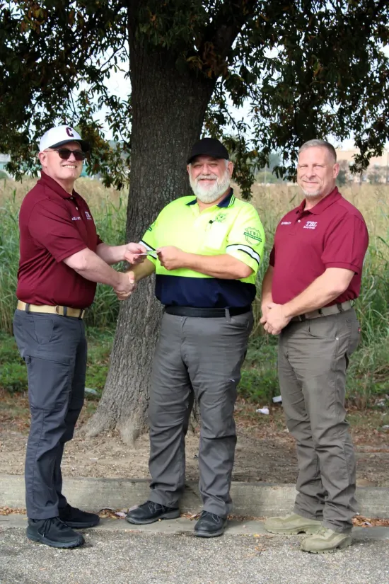 Three people stand outdoors near a tree, with the person on the left shaking hands with the person in the center.