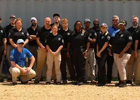 A group of people in matching uniforms stand outdoors against a corrugated metal wall for a team portrait.