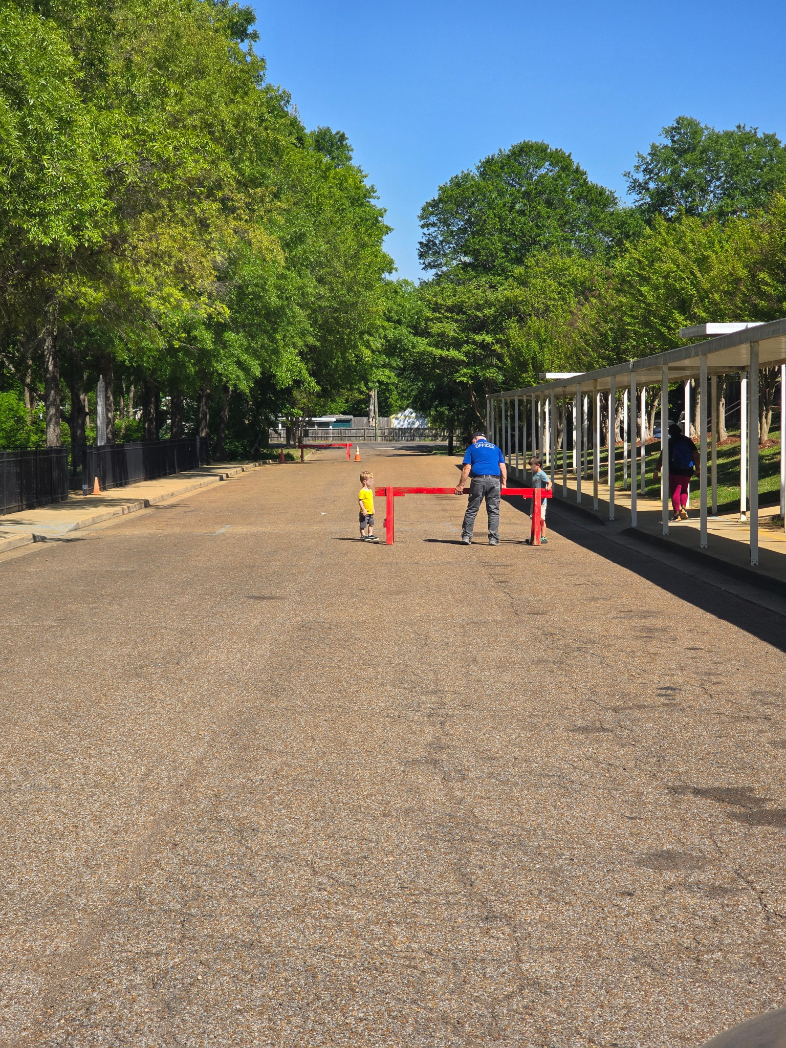 A person stands near a red barrier on a gravel path lined with trees and a white covered walkway.
