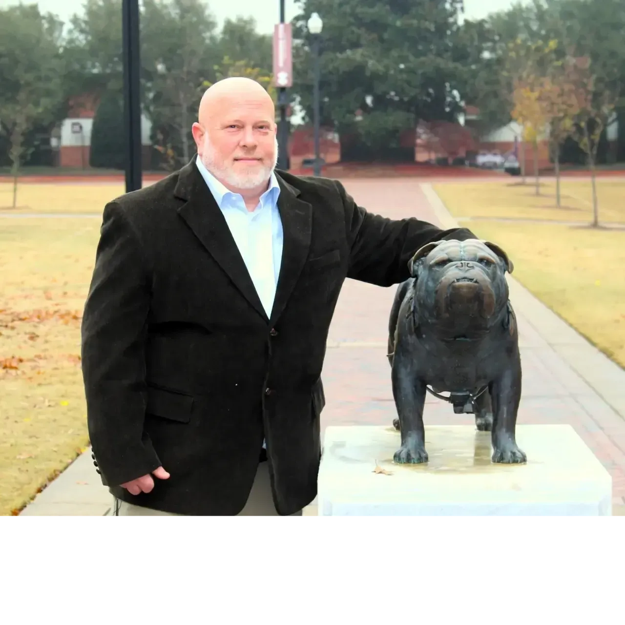 A person in a dark blazer stands outside on a campus path, resting a hand on a bronze bulldog statue.