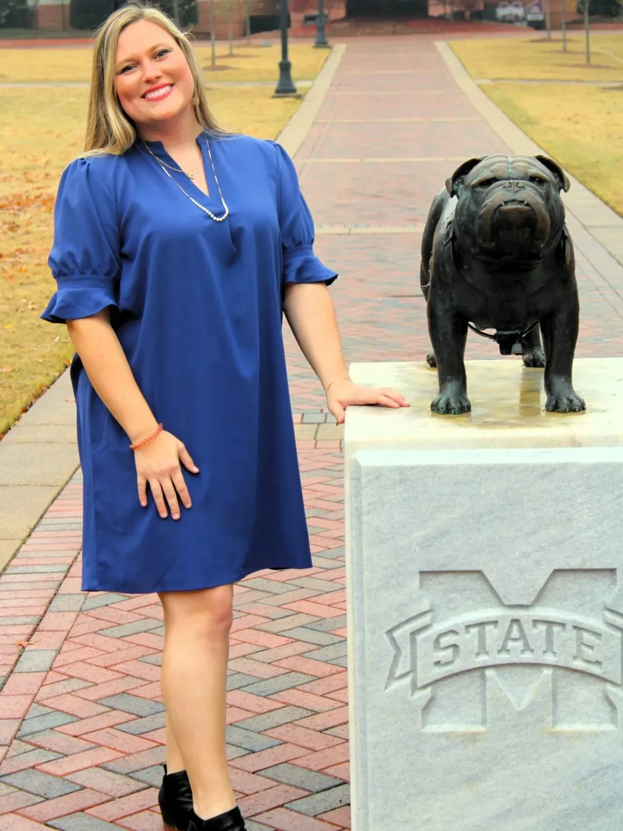 A person in a blue dress stands smiling next to a bronze bulldog statue on a Mississippi State University campus path.