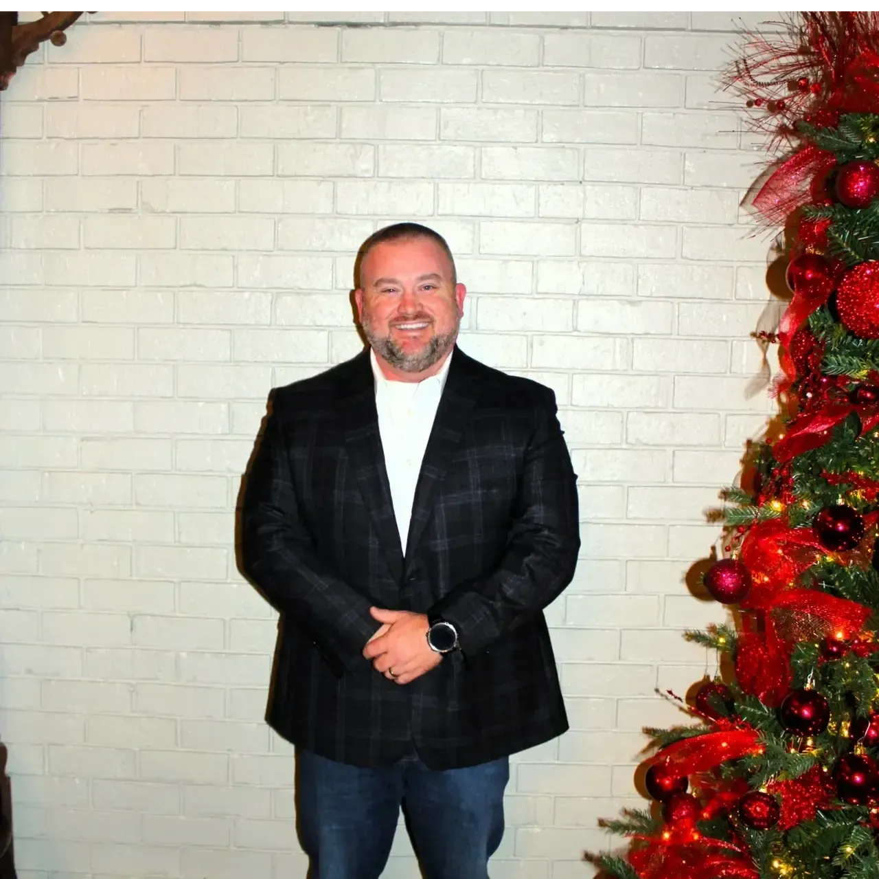 A smiling man stands with hands clasped in front of a white brick wall next to a decorated Christmas tree.