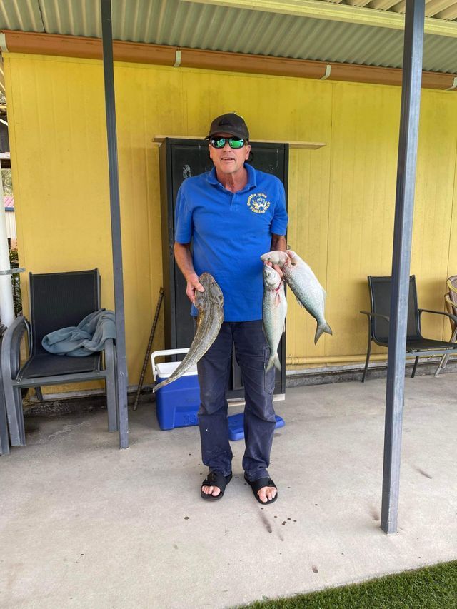 A Man In A Blue Shirt Is Holding Two Fish In Front Of A Yellow Building — Pacific Palms Bowling Club In Smiths Lake, NSW