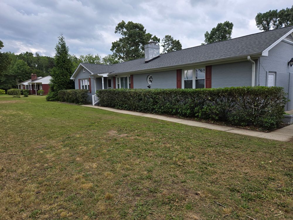 Gray house with a long hedge row and green lawn under a cloudy sky.