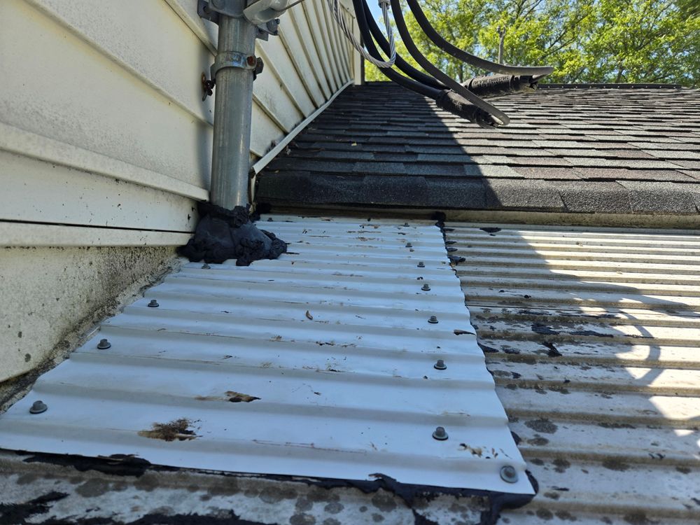 White siding and corrugated metal meet a shingled roof; black sealant applied around pipe and metal.