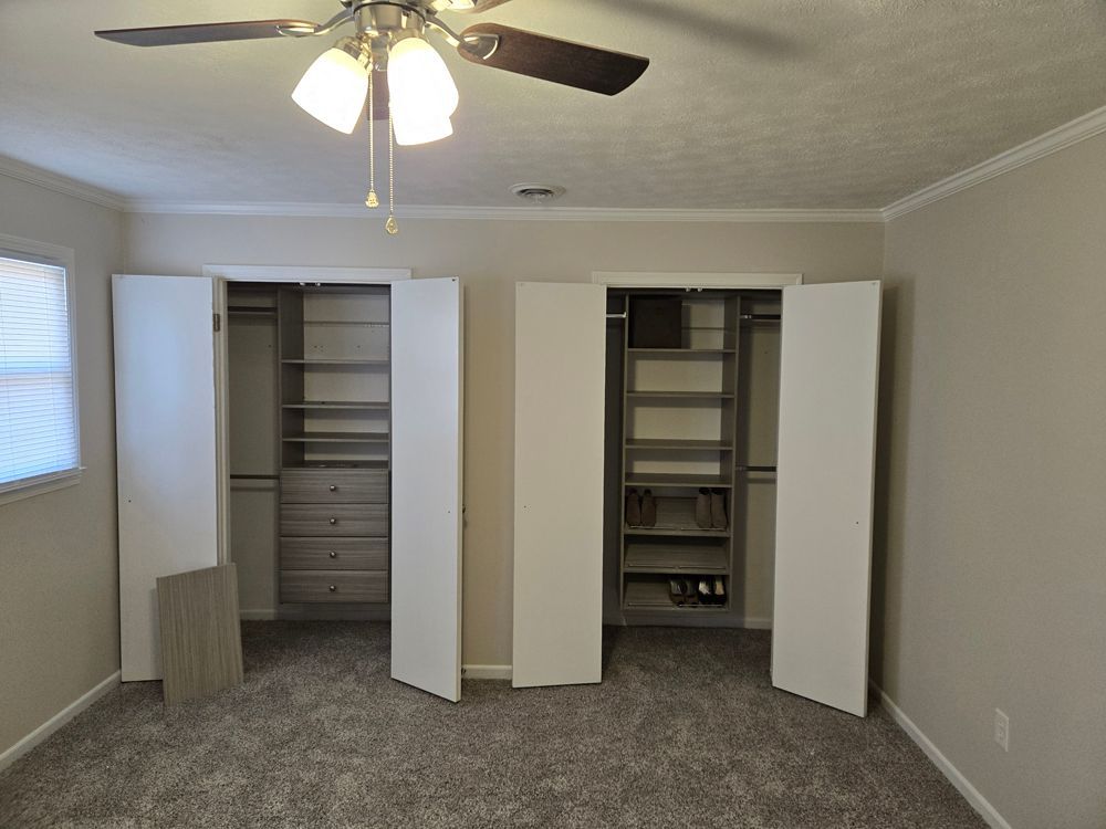 A bedroom with two open closets and organized shelves, light gray walls and carpet, and a ceiling fan.