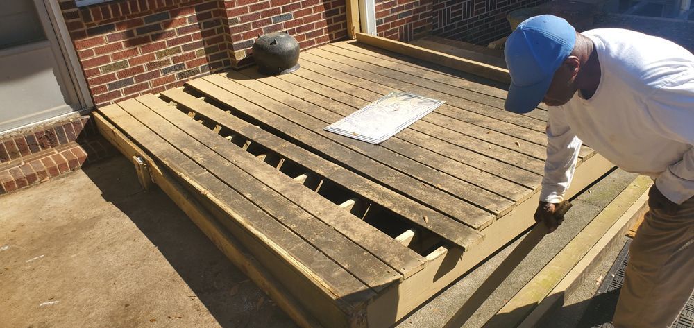 Man examines a deteriorating wooden deck with missing planks next to a brick wall.